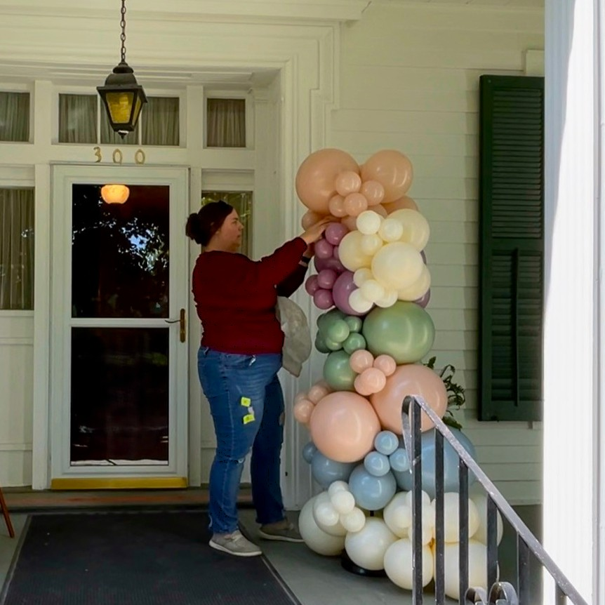 Balloon column in soft pastel tones being adjusted beside a porch entrance, featuring blush, mauve, sage, cream, and light blue balloons for a baby shower display.