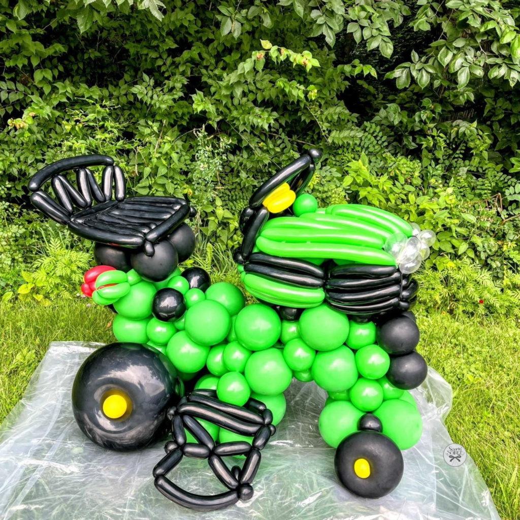 Green and black balloon sculpture shaped like a lawn mower, displayed outdoors on grass with detailed wheels and seat.