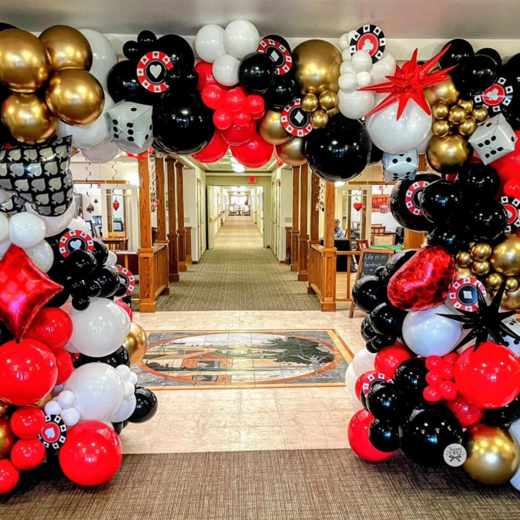 Red, black, white, and gold balloon arch with playing card, dice, and poker chip accents framing a hallway entrance for a casino-themed event.