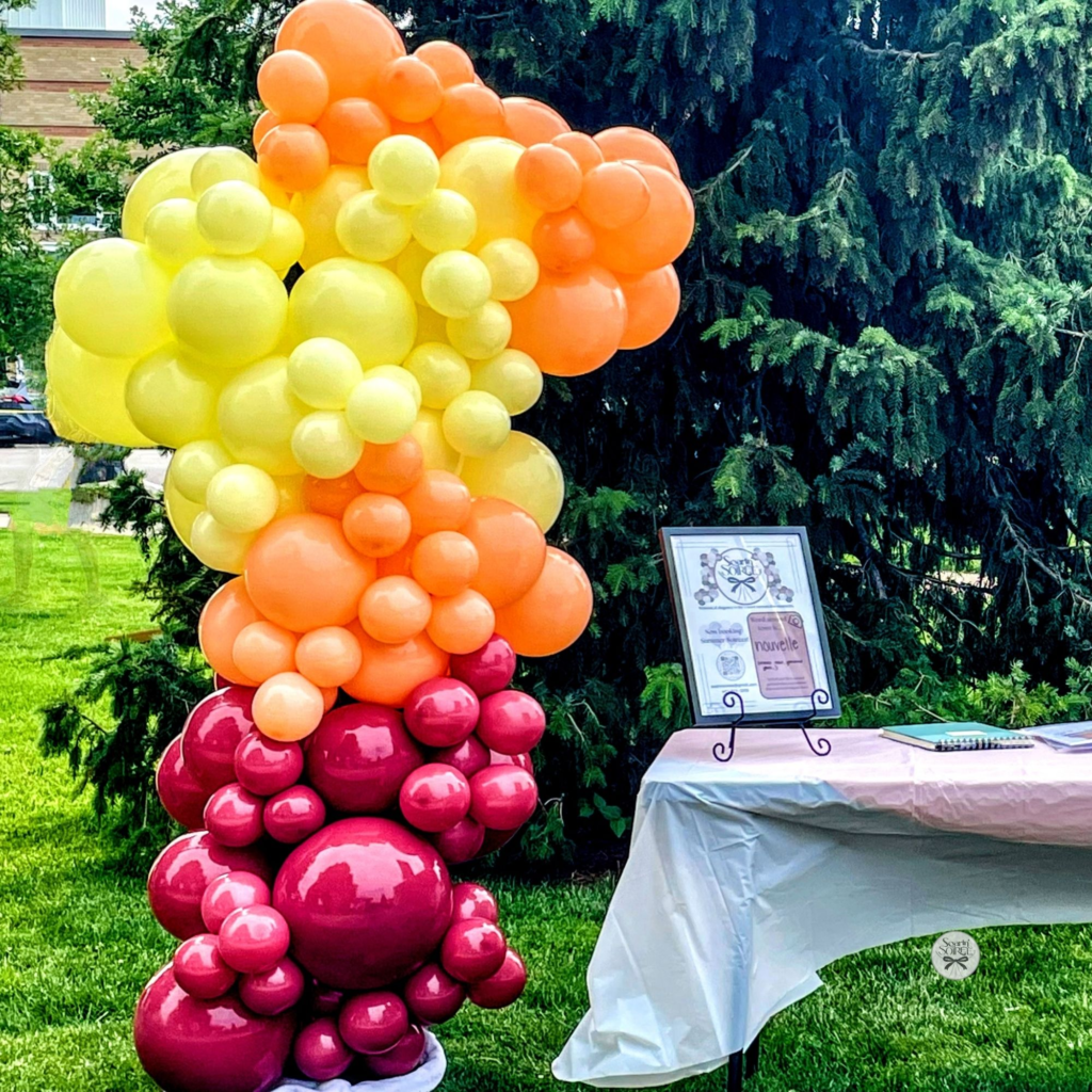 Vertical balloon column in yellow, orange, and deep berry tones displayed outdoors beside a table, creating a bright, welcoming accent for a community event.