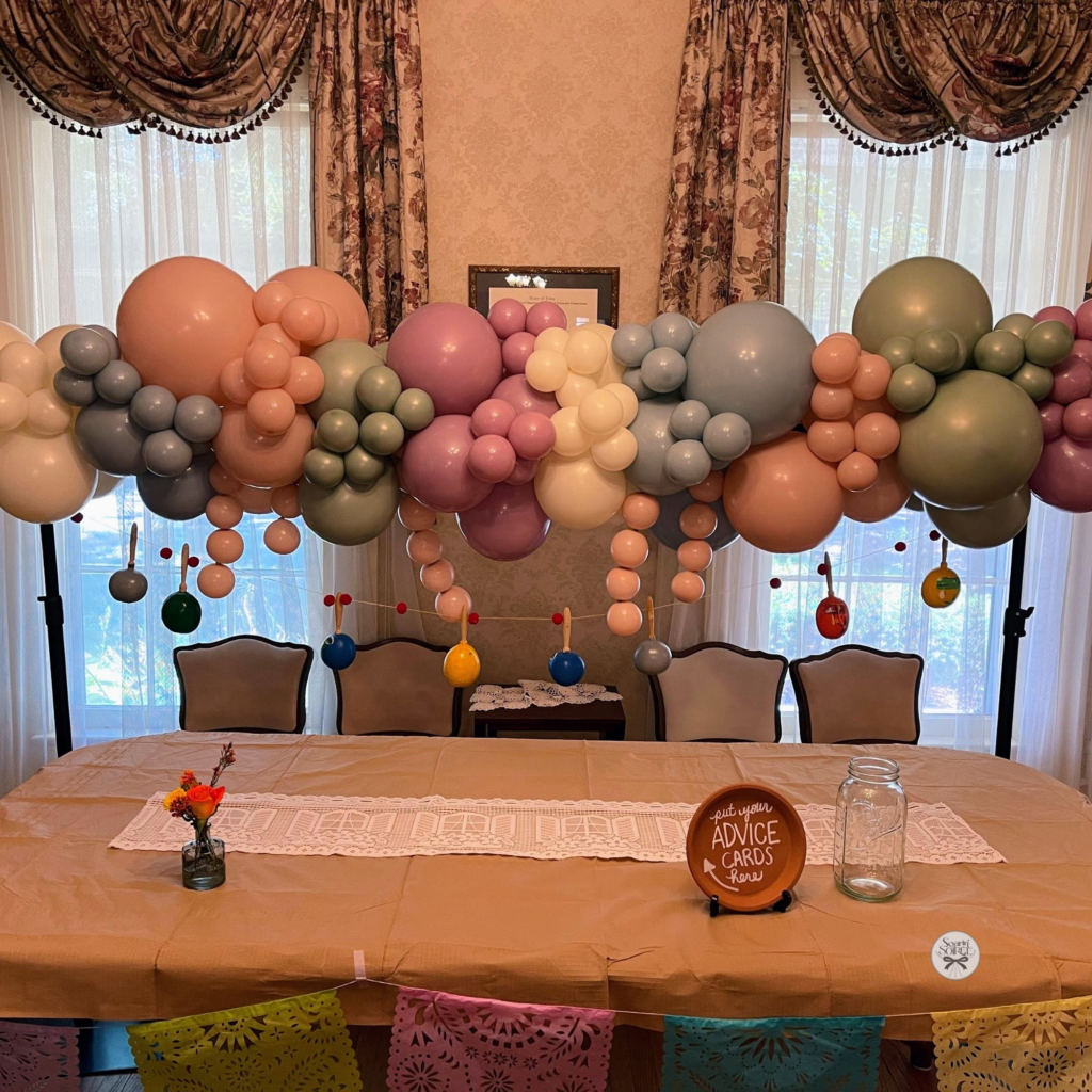 Pastel balloon garland in blush, sage, lavender, cream, and gray styled above a long table for a baby shower.