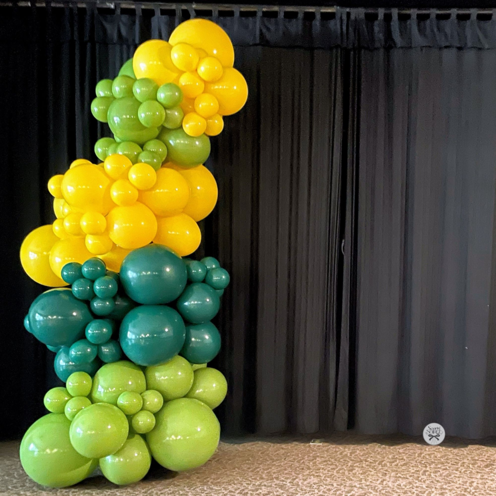 Vertical balloon column in shades of green, teal, and yellow set against a black curtain backdrop, creating a bold, modern event accent.