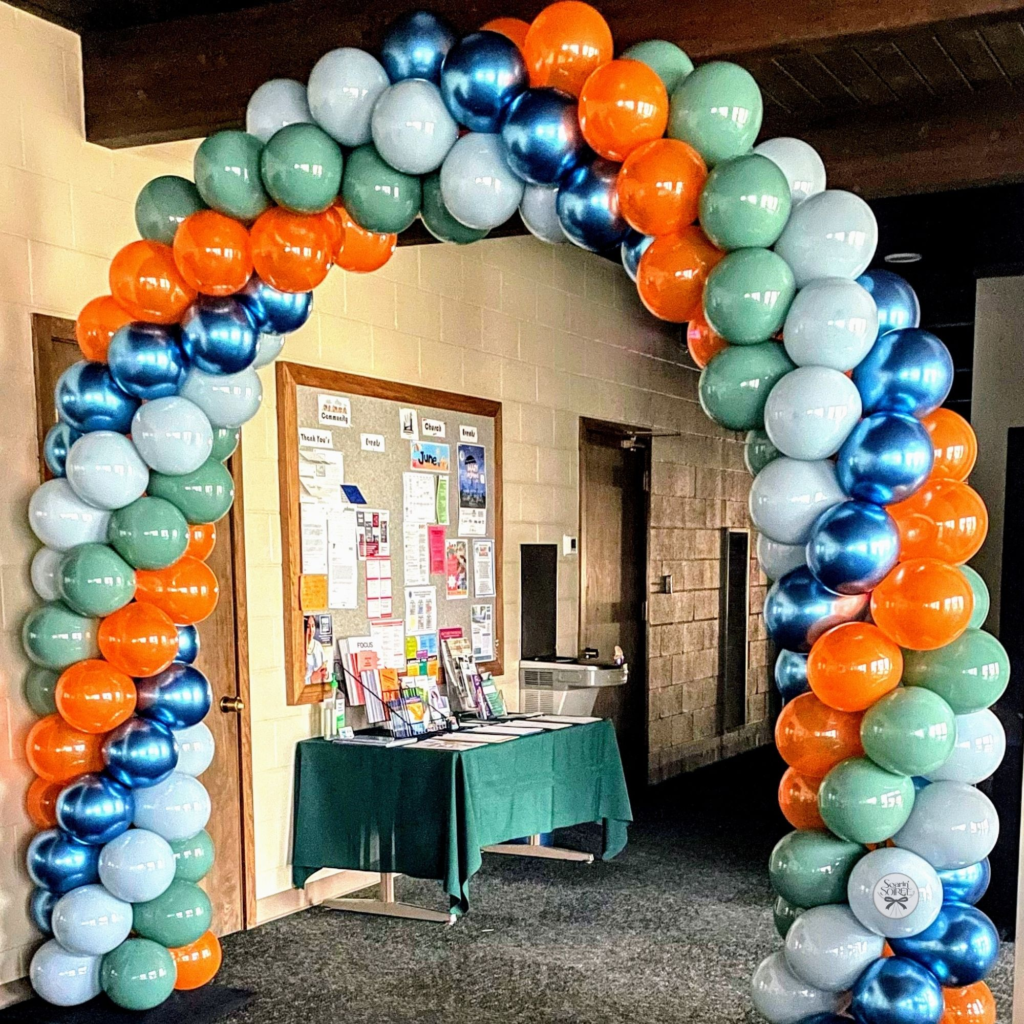 Balloon arch in teal, light blue, metallic blue, and orange framing an indoor entryway, creating a welcoming display for a church event.