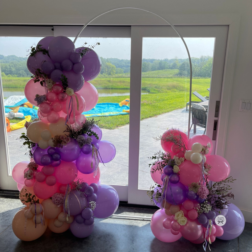 Pastel balloon arch with pink, lavender, and peach balloons accented with florals, framing a bright indoor doorway.