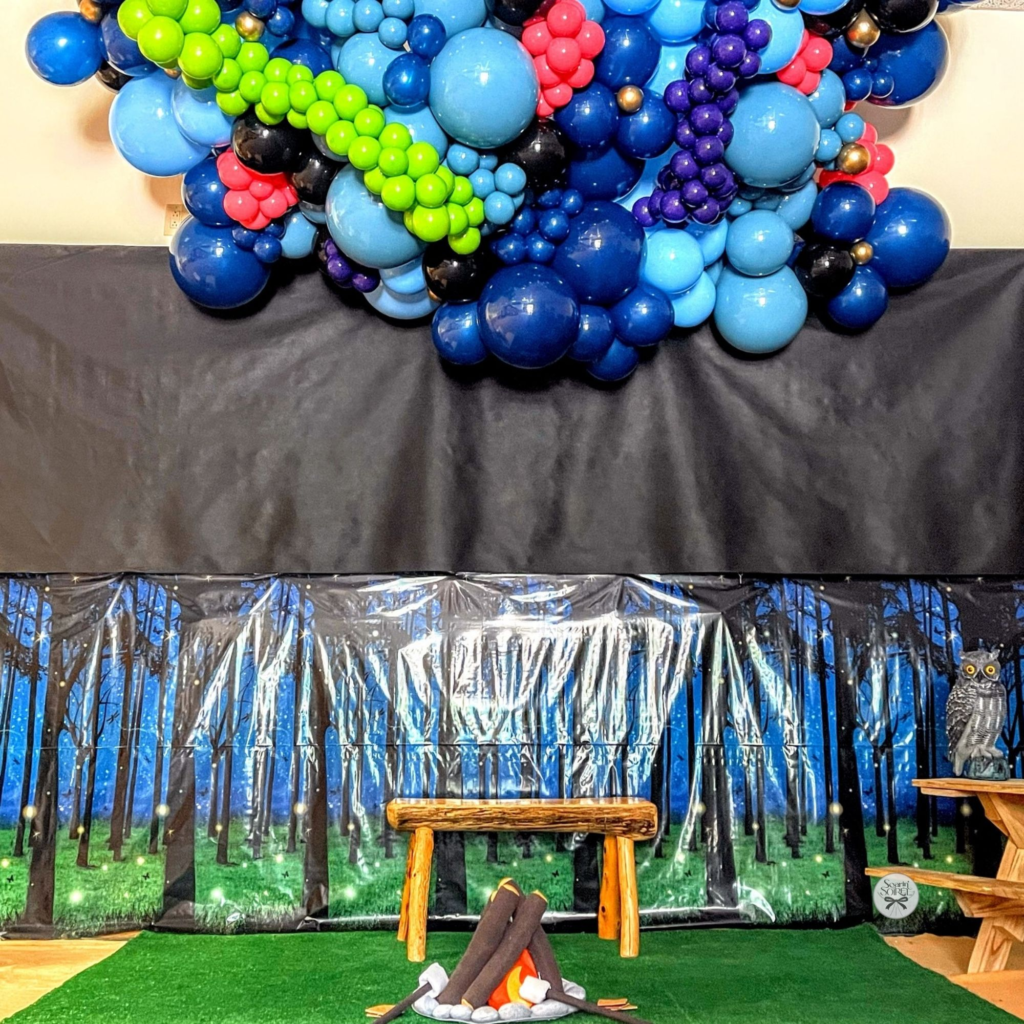 Blue, green, purple, and black balloon installation designed to represent the northern lights above a forest-themed backdrop with a campfire scene.
