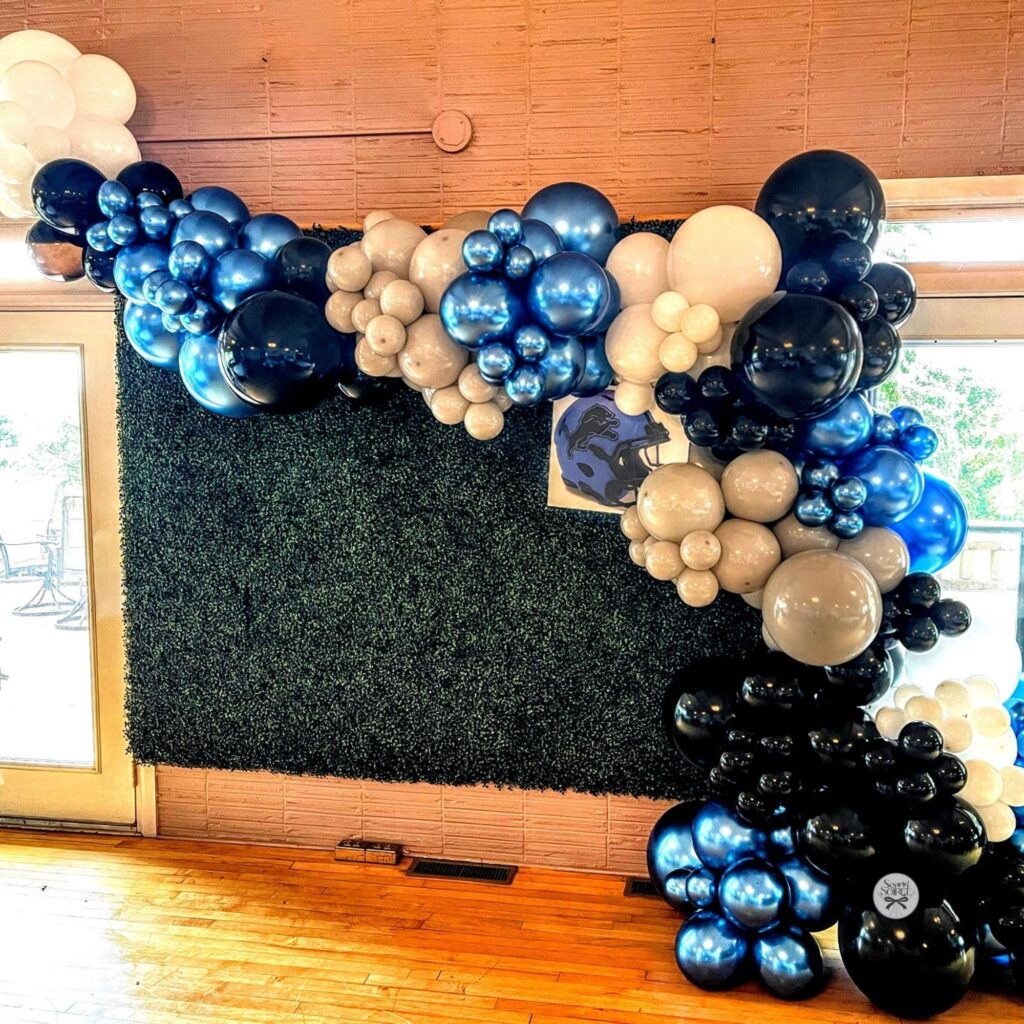 Blue, black, white, and silver balloon garland framing a green hedge backdrop indoors, creating a bold photo display for a community event.
