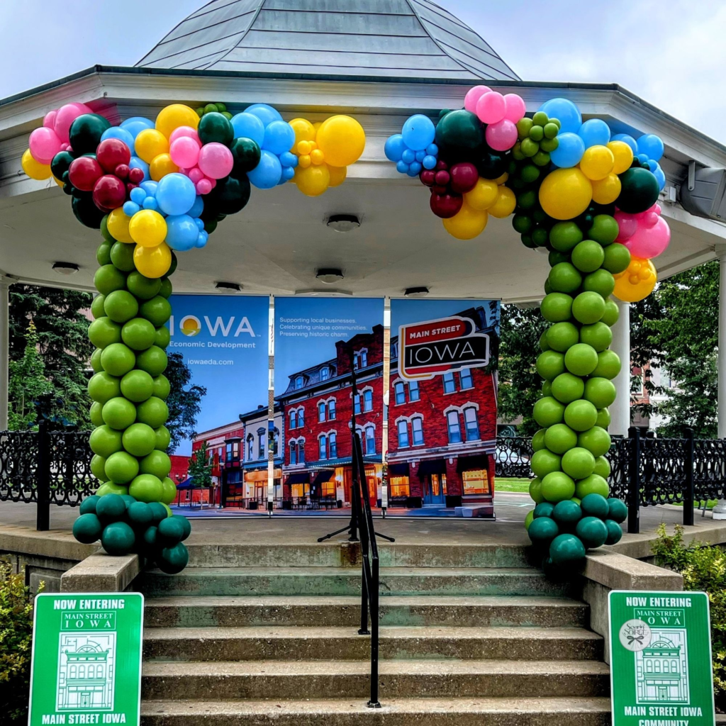 Balloon arch styled as a tree with a green trunk and multicolored canopy, installed on a gazebo stage for a community event.