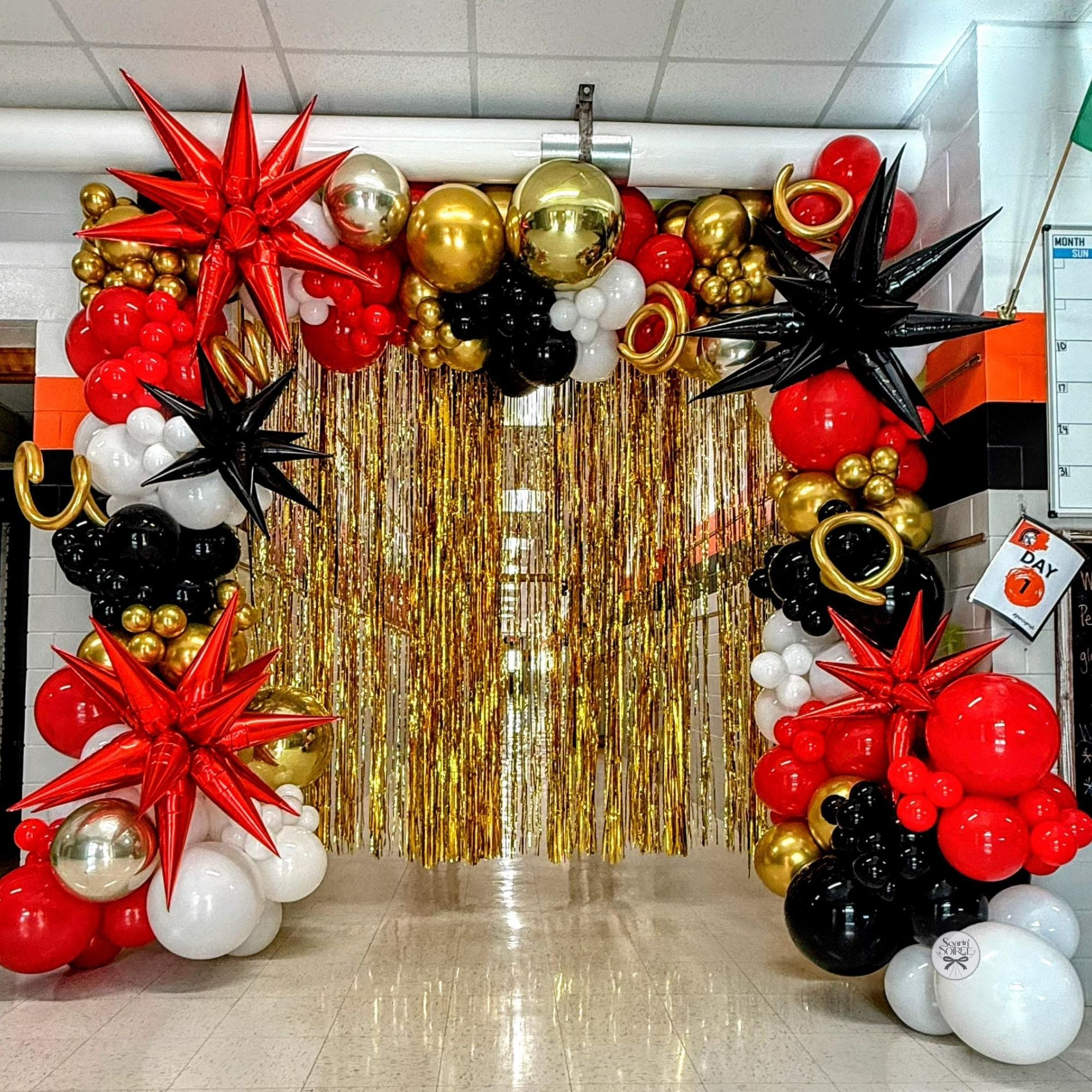 Red, black, white, and gold balloon arch with starburst accents framing a gold fringe backdrop in a school hallway, creating a bold Hollywood-themed photo area.