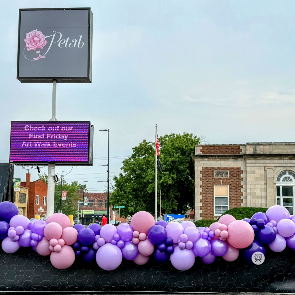Lavender, purple, and blush balloon garland styled along an outdoor planter wall in a downtown streetscape.