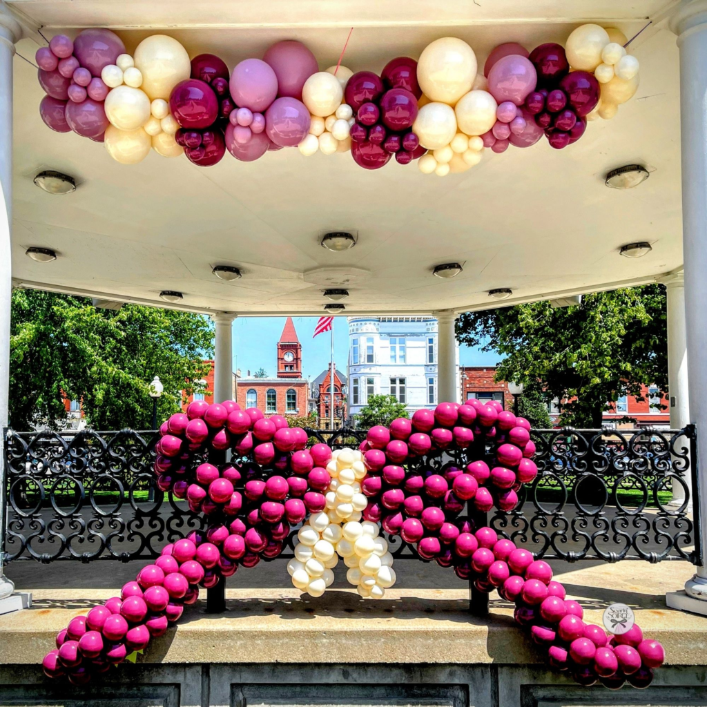 Large magenta balloon bow with a cream center displayed under a gazebo, paired with a coordinating pastel balloon garland overhead in a town square.