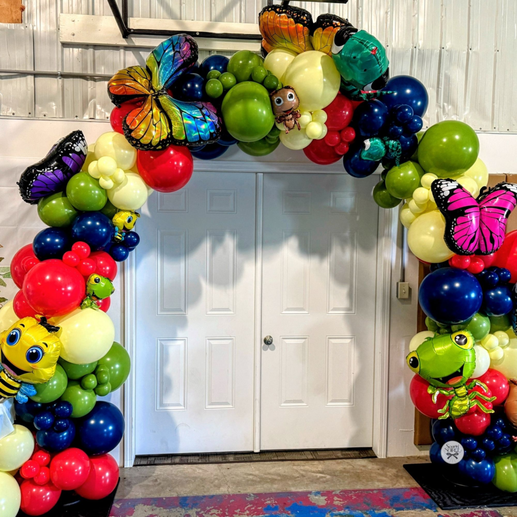 Colorful balloon arch with butterfly and bug accents framing double doors, creating a playful garden-themed entrance.