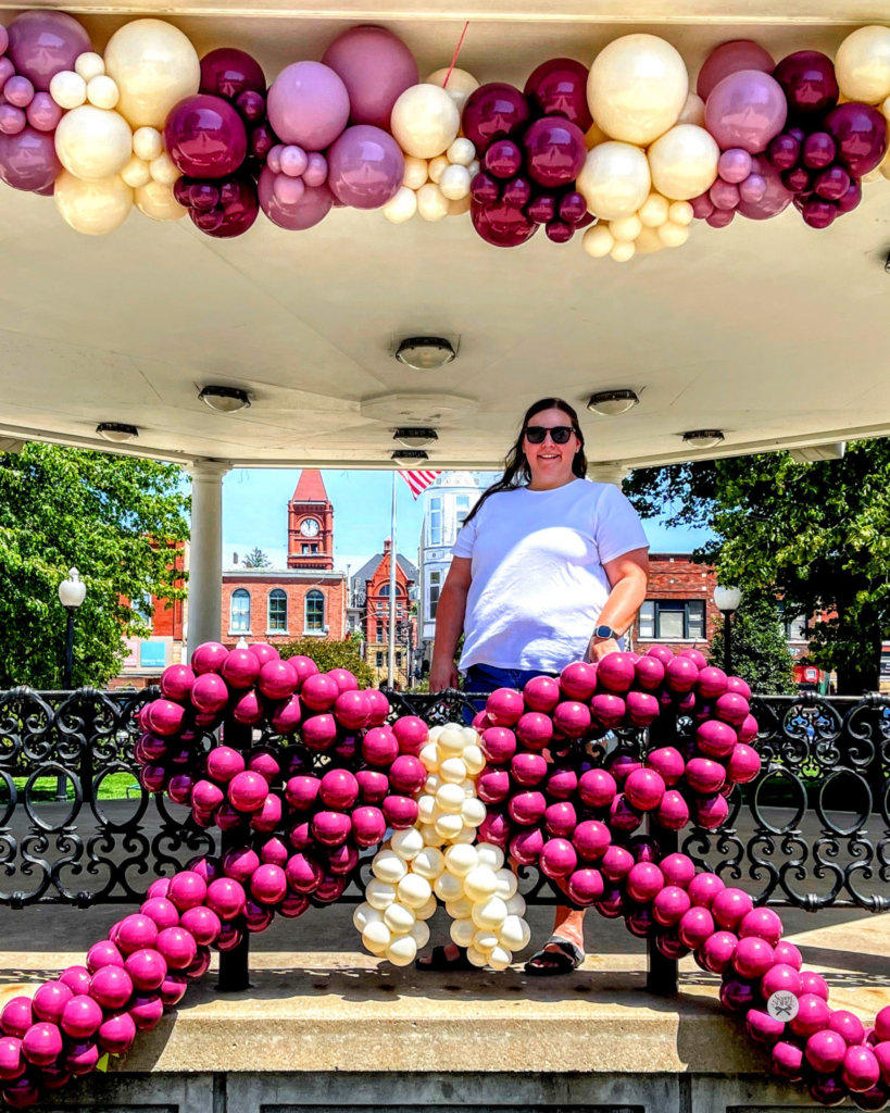 Large magenta balloon bow with a cream center displayed under a gazebo, paired with a coordinating pastel balloon garland overhead in a town square.